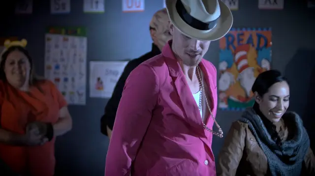 A dancer in a pink blazer and fedora leads a group dance in a classroom-style set.